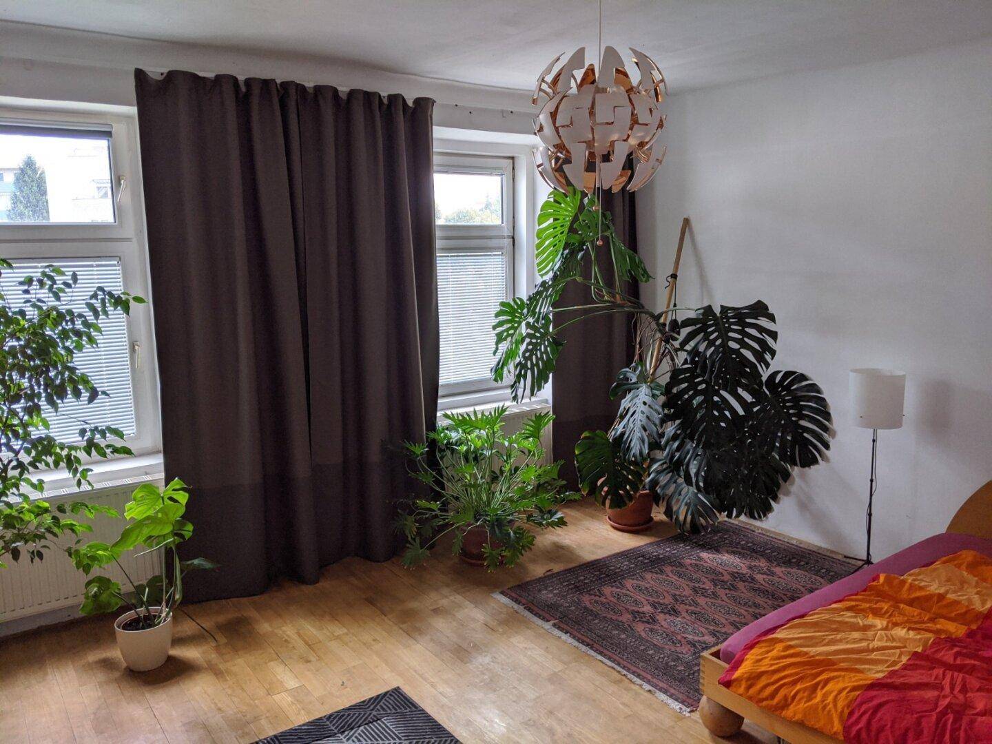 Helles Schlafzimmer mit großen Pflanzen, Holzboden und einem farbenfrohen Bett.