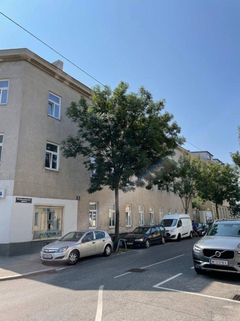 Seitenansicht des Gebäudes mit Bäumen und parkenden Fahrzeugen entlang der Straße unter blauem Himmel.