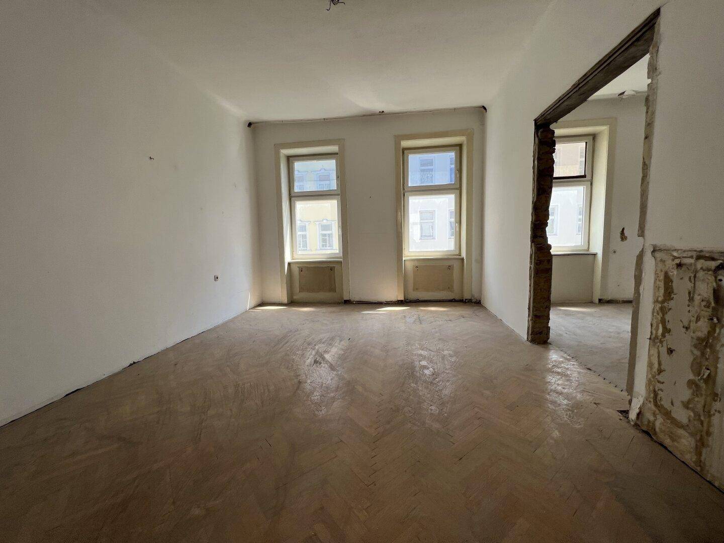 Heller Raum mit Fischgrätparkett und einem offenen Durchgang zu einem weiteren Zimmer.