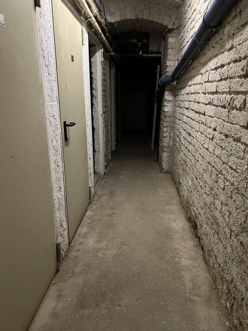 Langer, dunkler Kellerflur mit unverputzten Ziegelwänden und sichtbaren Rohren.