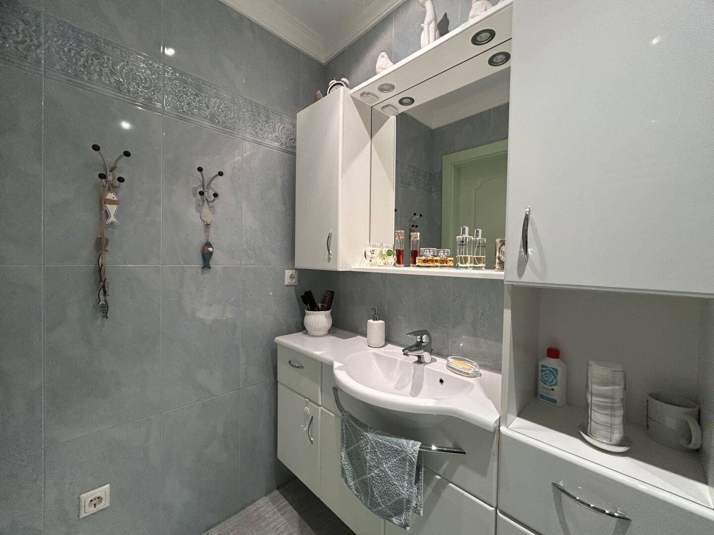 Badezimmer mit Waschbecken, Spiegelschrank und weißen Unterschränken auf grauen Fliesen.