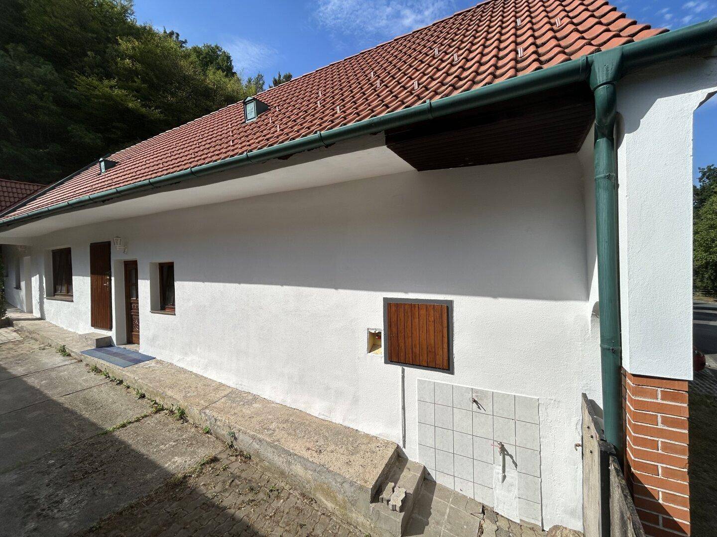 Seitenansicht des Hauses mit weißer Fassade, Regenrinne und einem kleinen gefliesten Bereich an der Wand.