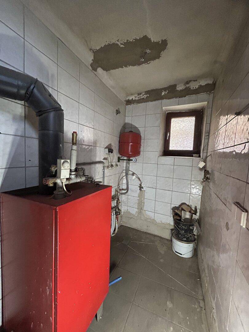 Heizungsraum mit rotem Boiler, sichtbaren Rohren und einem kleinen Fenster, in renovierungsbedürftigem Zustand.