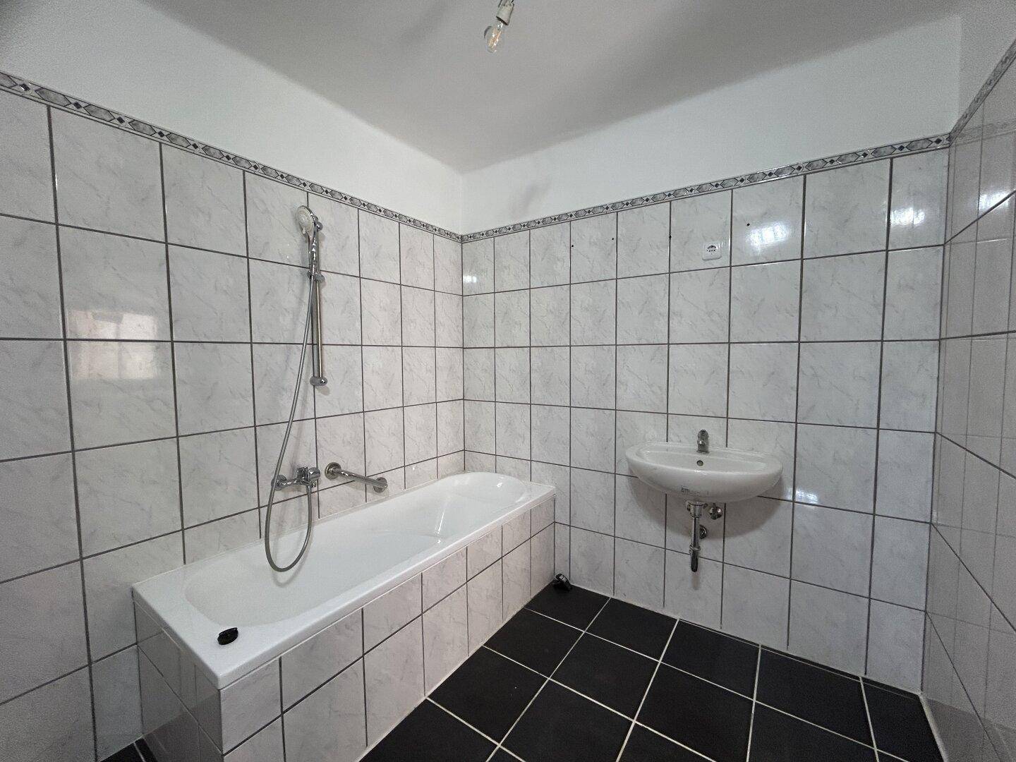 Badezimmer mit weißen Wandfliesen, schwarzen Bodenfliesen, Badewanne mit Duschfunktion und Waschbecken.
