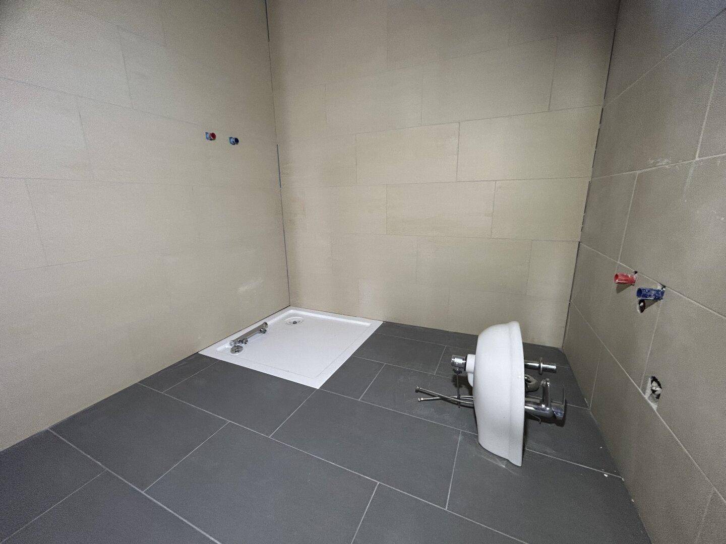 Unfertiges Badezimmer mit grauen Fliesen, Duschwanne und vorbereiteten Anschlüssen.