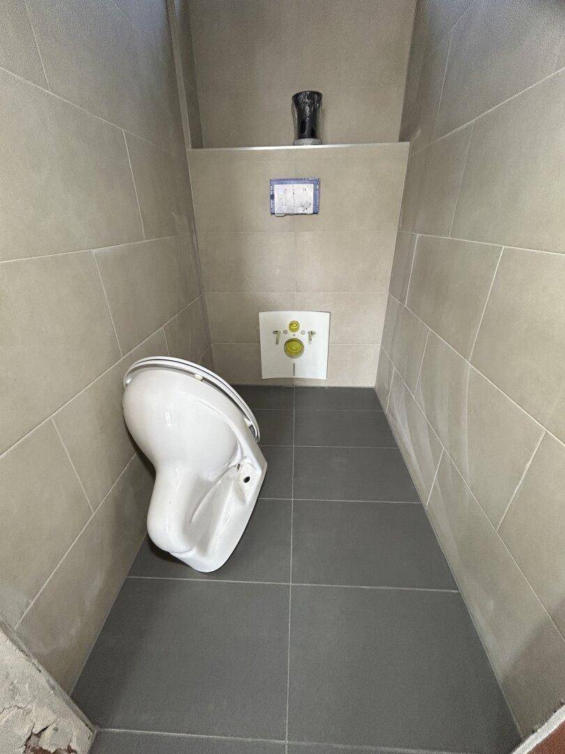 Unfertiges WC mit grauen Bodenfliesen und vorbereiteter Wandmontage für die Toilette.