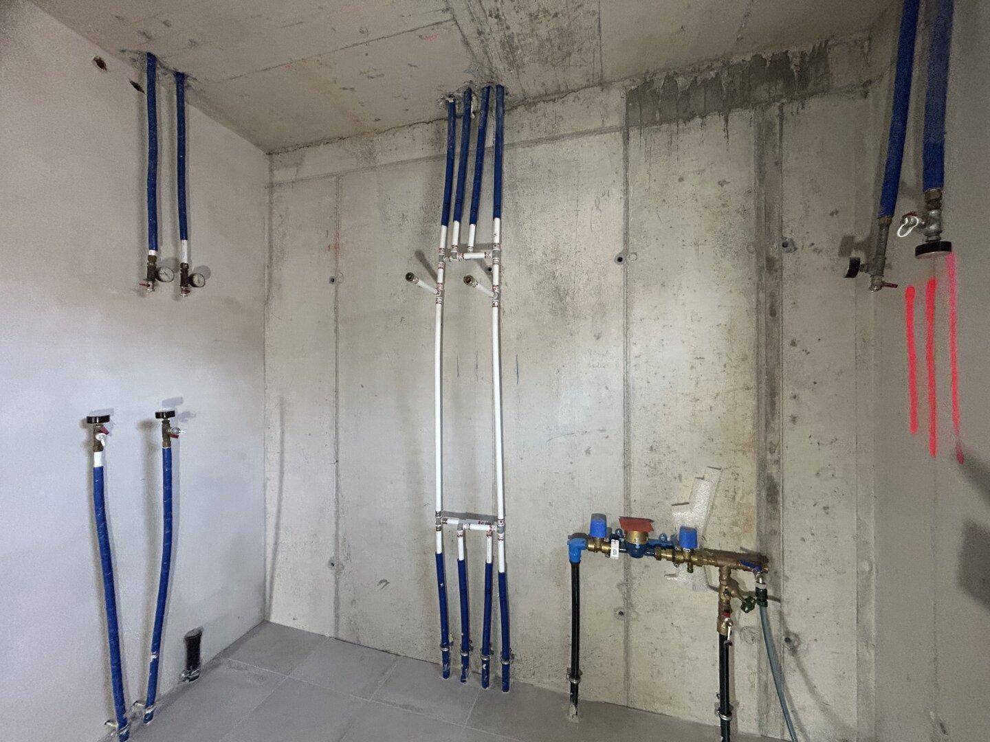 Technikraum im Rohbau mit sichtbaren Wasserleitungen und Betonwänden.