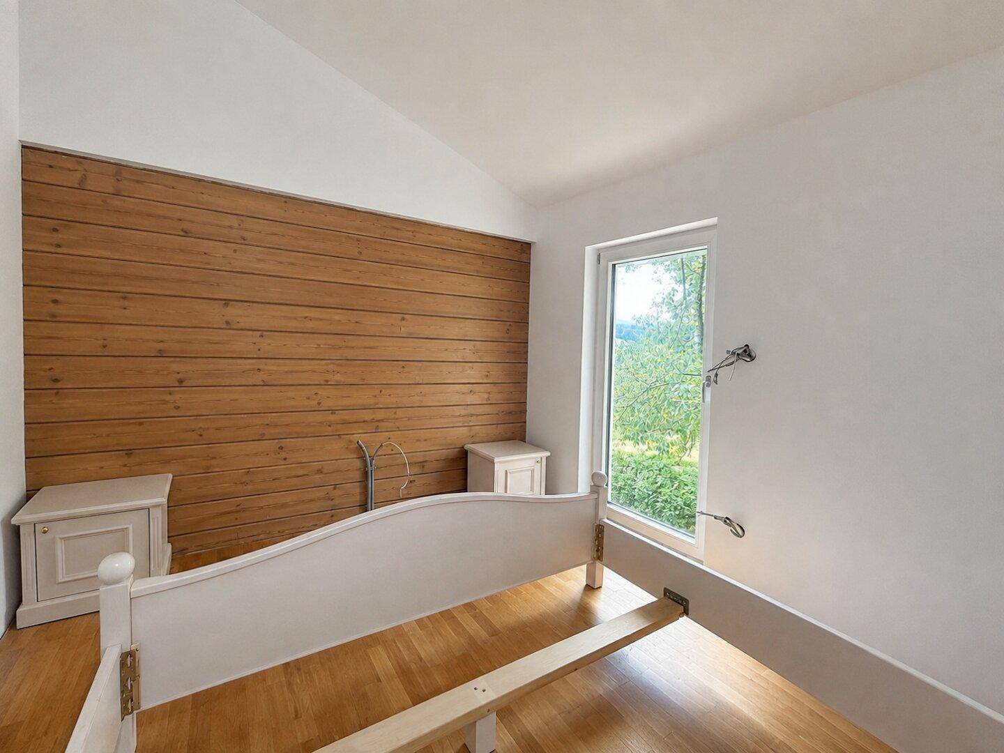 Schlafzimmer mit Holzwand, Parkettboden und Fenster mit Blick ins Grüne.