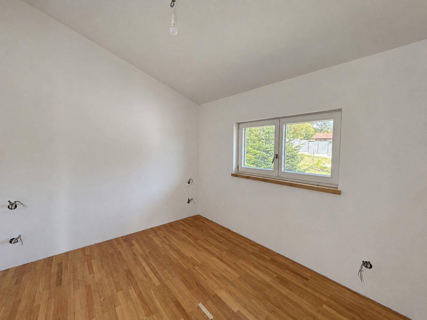 Geräumiges Zimmer mit Parkettboden und großem Fenster mit Blick ins Grüne.