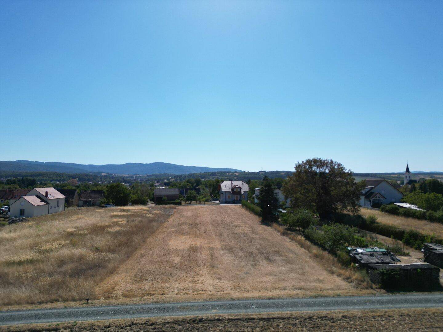 Panoramablick über weite Felder, Wohnhäuser und eine hügelige Landschaft mit Bergen im Hintergrund.