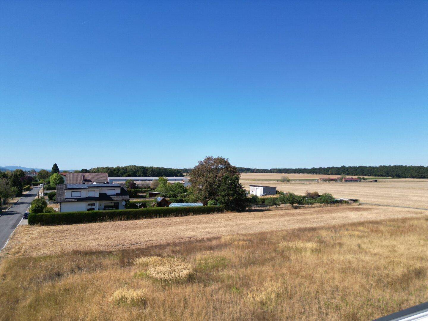 Ländliche Szenerie mit trockenen Feldern, vereinzelten Häusern und einem Waldstreifen am Horizont.