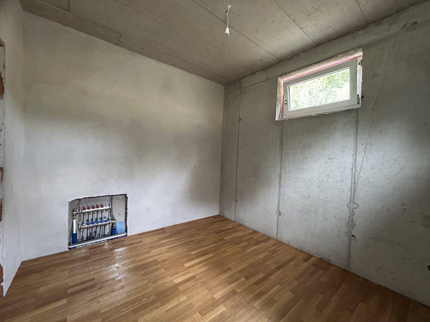 Unfertiger Raum mit Parkettboden, kleinem Fenster und sichtbaren Installationen für die Heizung.