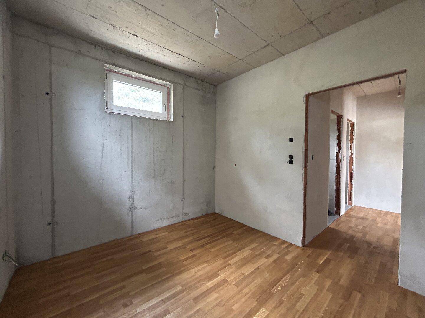 Rohbau-Zimmer mit Parkettboden, kleinem Fenster und Zugang zu weiteren unfertigen Bereichen.
