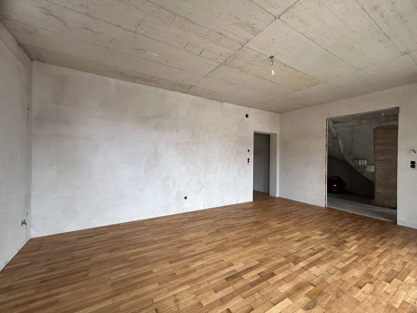 Unfertiger Wohnraum mit neu verlegtem Parkettboden und unverputzten Betonwänden, bereit für den Ausbau.