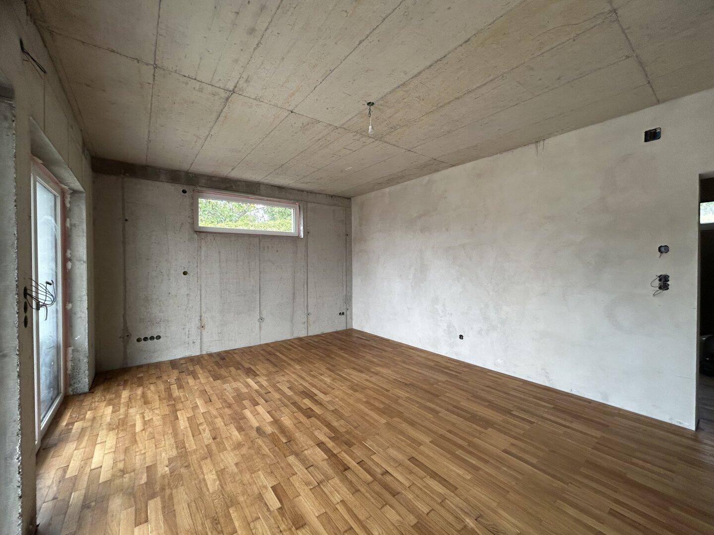 Unfertiger Raum mit Sichtbetondecke, Parkettboden und großen Fenstern für viel Licht.