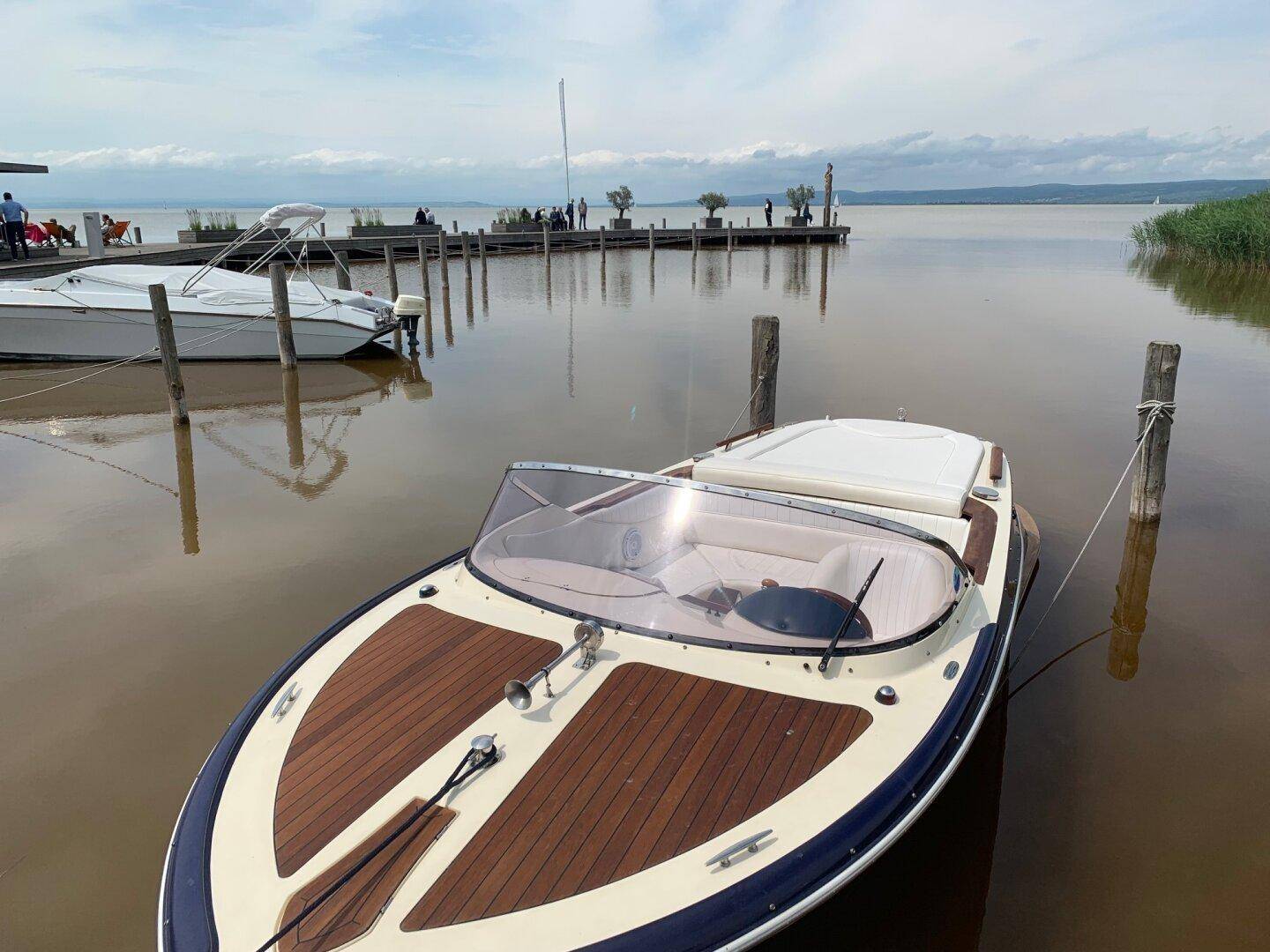 Ein elegantes Motorboot liegt an einem Steg auf einem ruhigen See unter bewölktem Himmel.