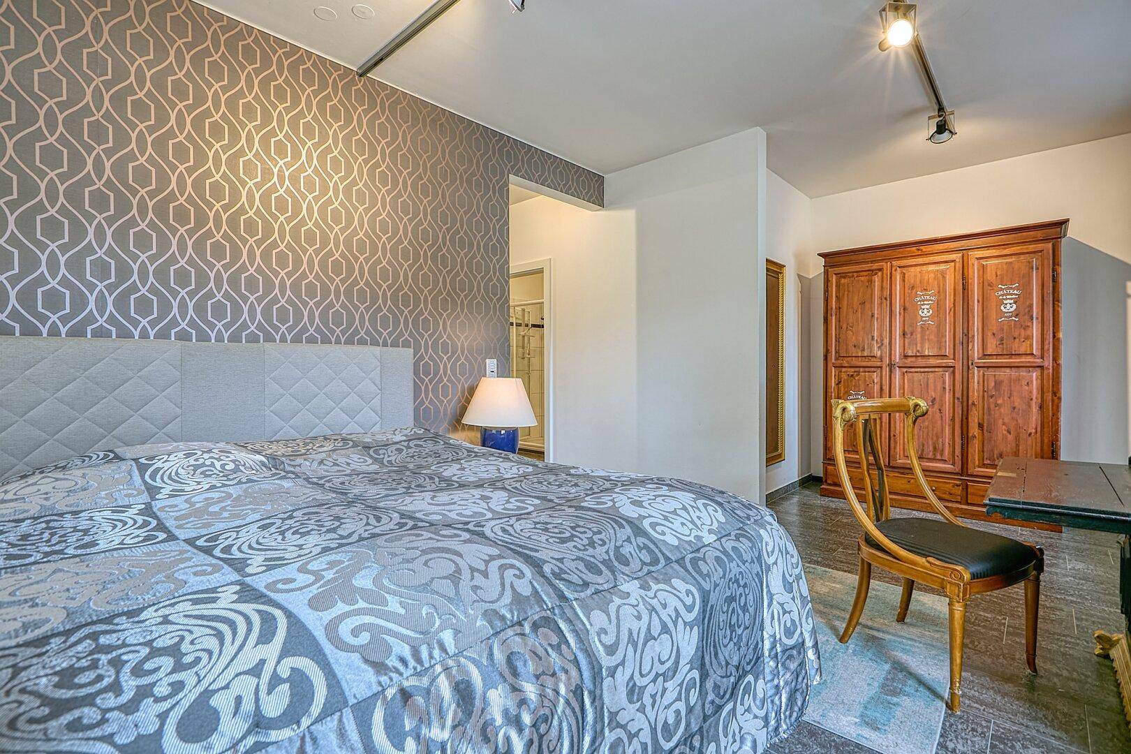 Schlafzimmer mit großem Bett, gemusterter Tapete und einem traditionellen Holzschrank.