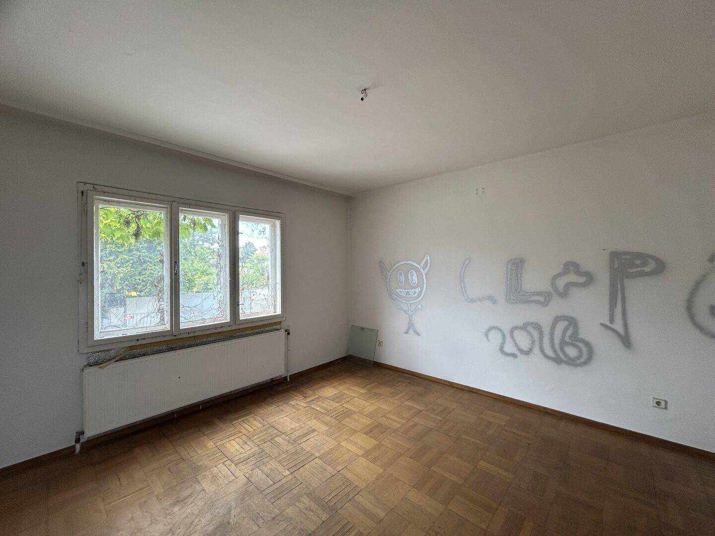 Raum mit Fischgrätparkett, Fenster und Graffiti, das auf den Zustand der Wände hinweist.
