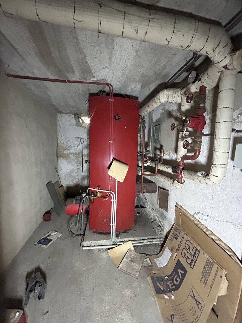 Kellerraum mit großem roten Warmwasserspeicher und sichtbaren Rohren, die Wände sind aus Beton.
