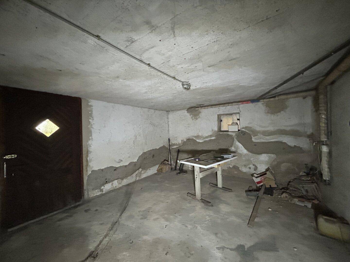 Leerer Kellerraum mit Betonboden und -wänden, einem Tisch und einer Holztür.
