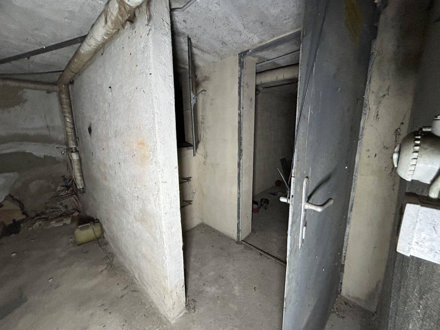 Unfertiger Kellerraum mit Betonwänden, sichtbaren Rohren und einer robusten Metalltür, ideal als Lagerfläche.