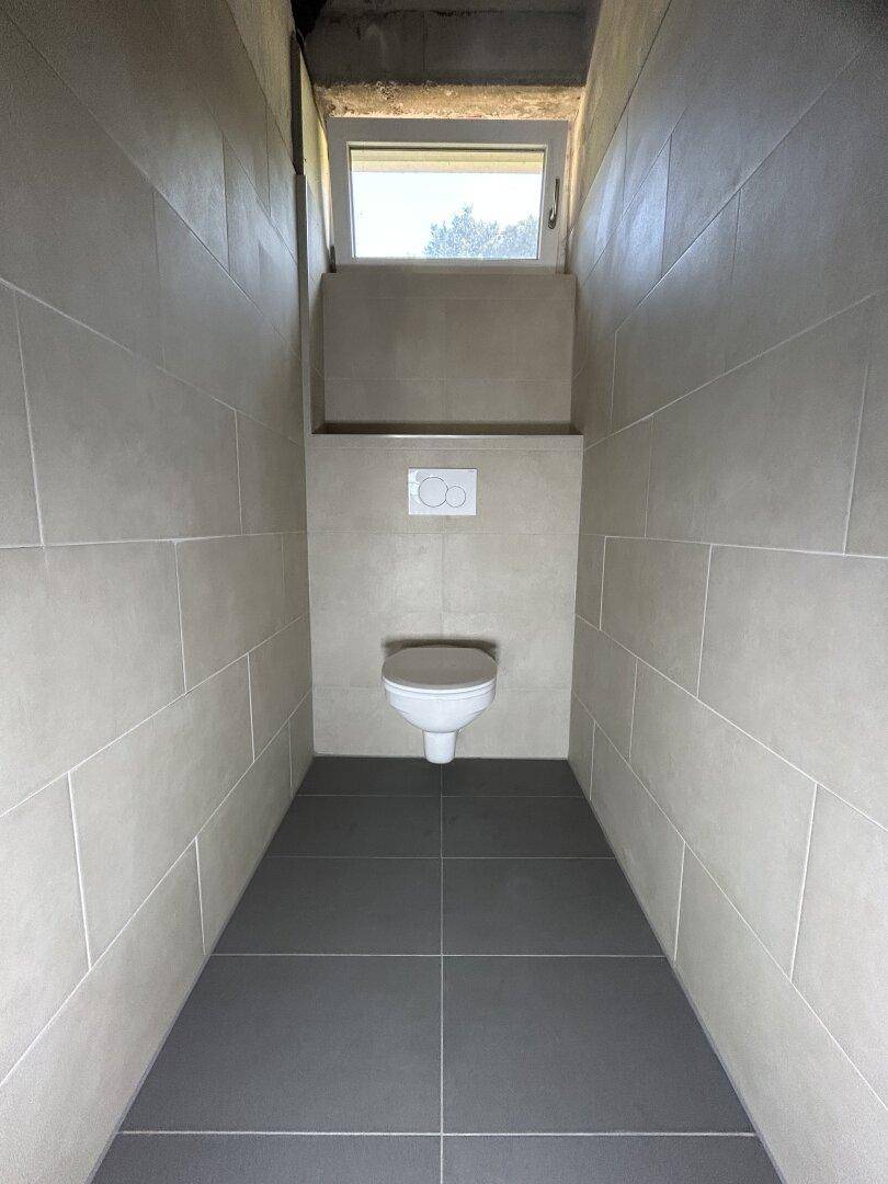 Modern gefliestes WC mit wandhängender Toilette und kleinem Fenster für natürliche Belüftung.