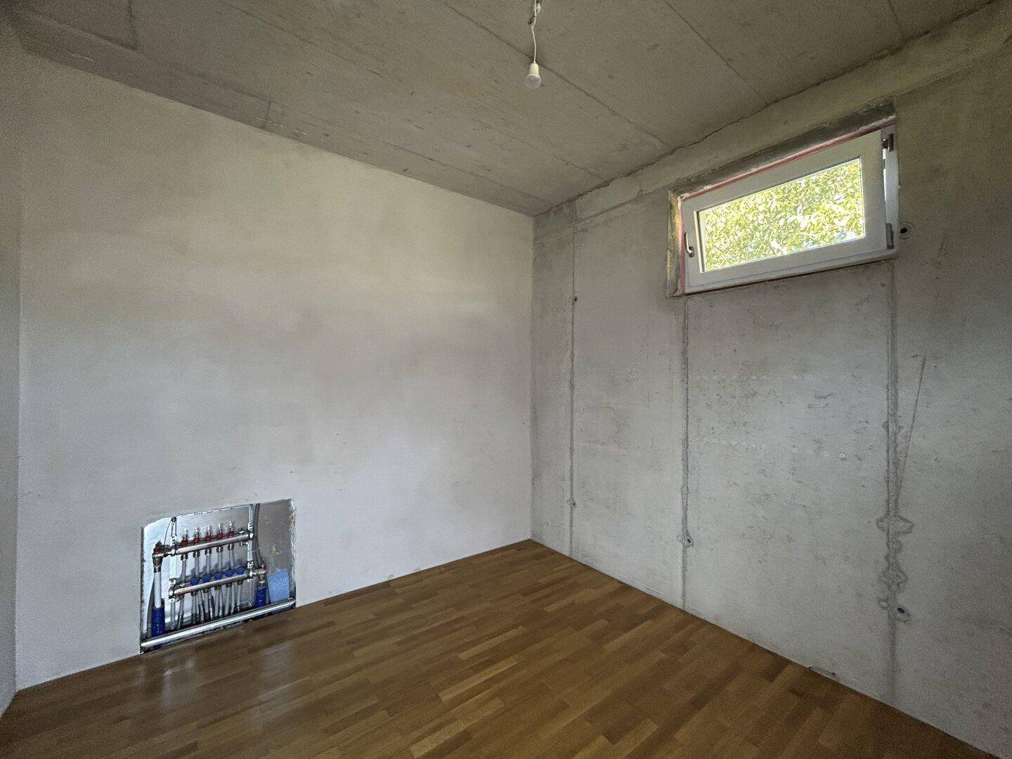 Unfertiger Raum mit Parkettboden, kleinem Fenster und sichtbaren Installationen für die Heizung.