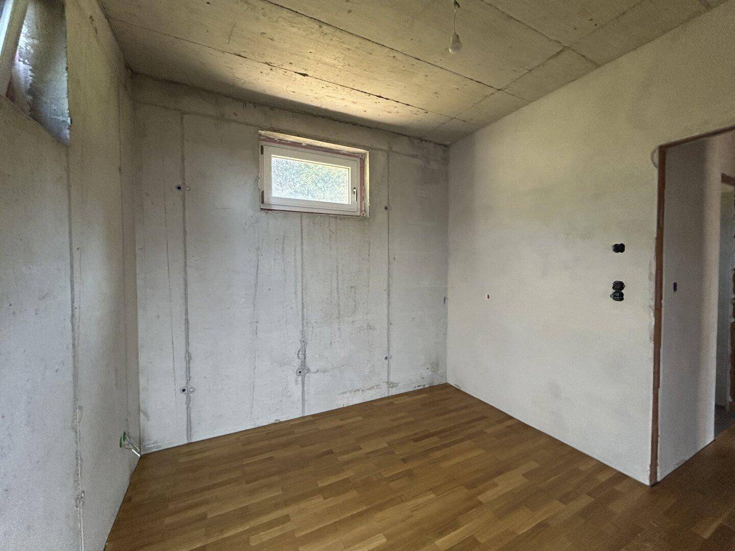 Rohbau-Zimmer mit Parkettboden, kleinem Fenster und Zugang zu weiteren unfertigen Bereichen.