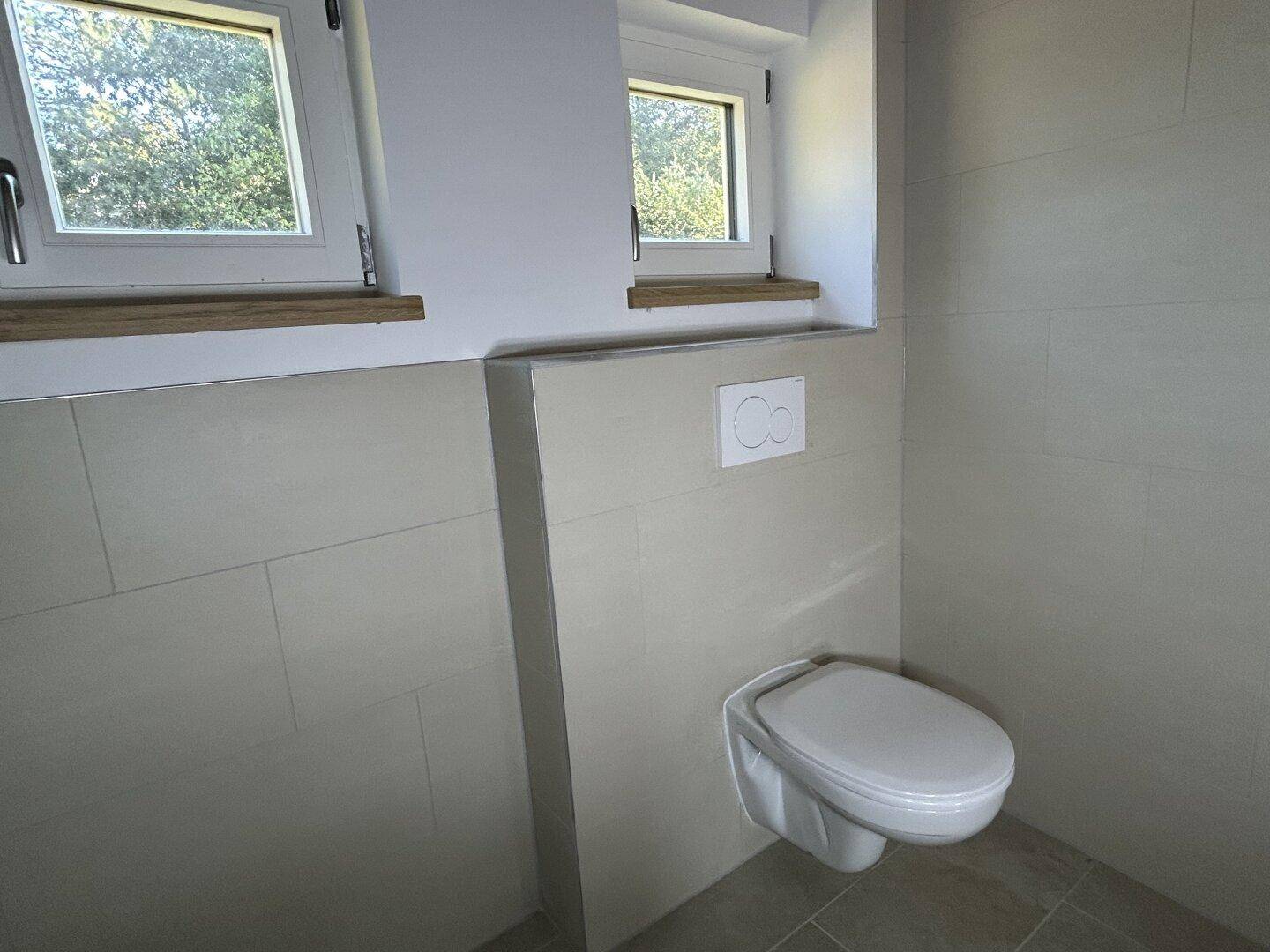 Helles WC mit wandhängender Toilette und zwei Fenstern, modern gefliest in neutralen Tönen.