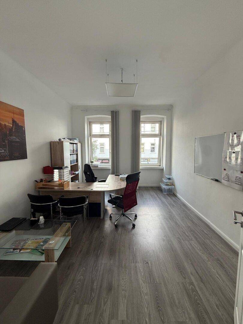 Geräumiges Büro mit grauem Laminatboden, Schreibtisch und Regalen, gut beleuchtet durch zwei Fenster.