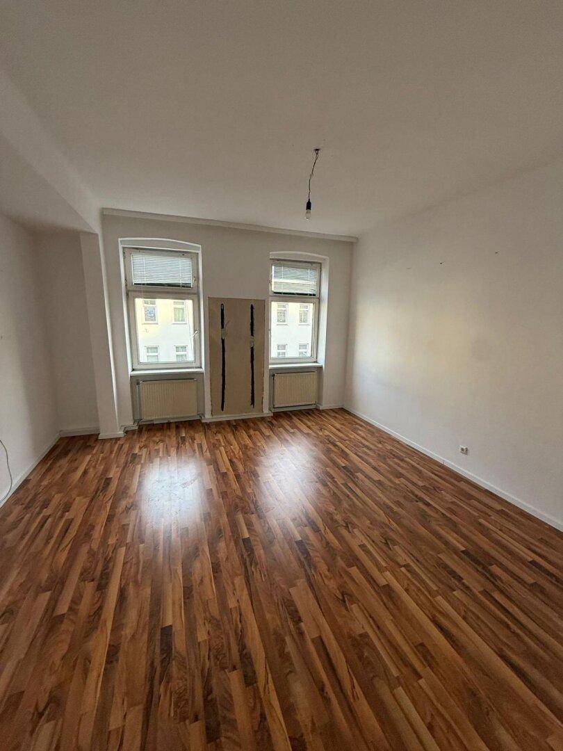 Helles, leeres Zimmer mit Laminatboden und zwei Fenstern, bereit zur individuellen Gestaltung.