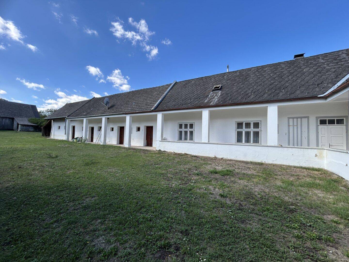 Lange Fassade eines ländlichen Gebäudes mit mehreren Türen und Fenstern, umgeben von Grünflächen.