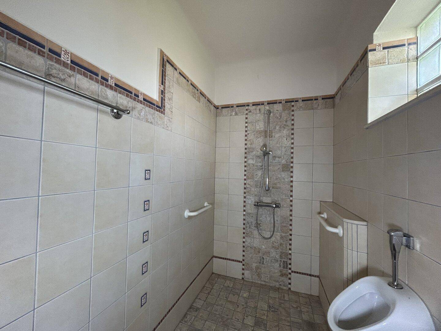 Duschbereich im Badezimmer mit Fliesen, Haltegriffen und einem Urinal.