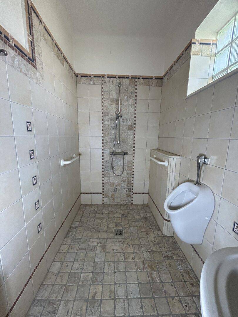 Geräumiger Duschbereich mit Fliesenboden, Haltegriffen und einem Urinal.
