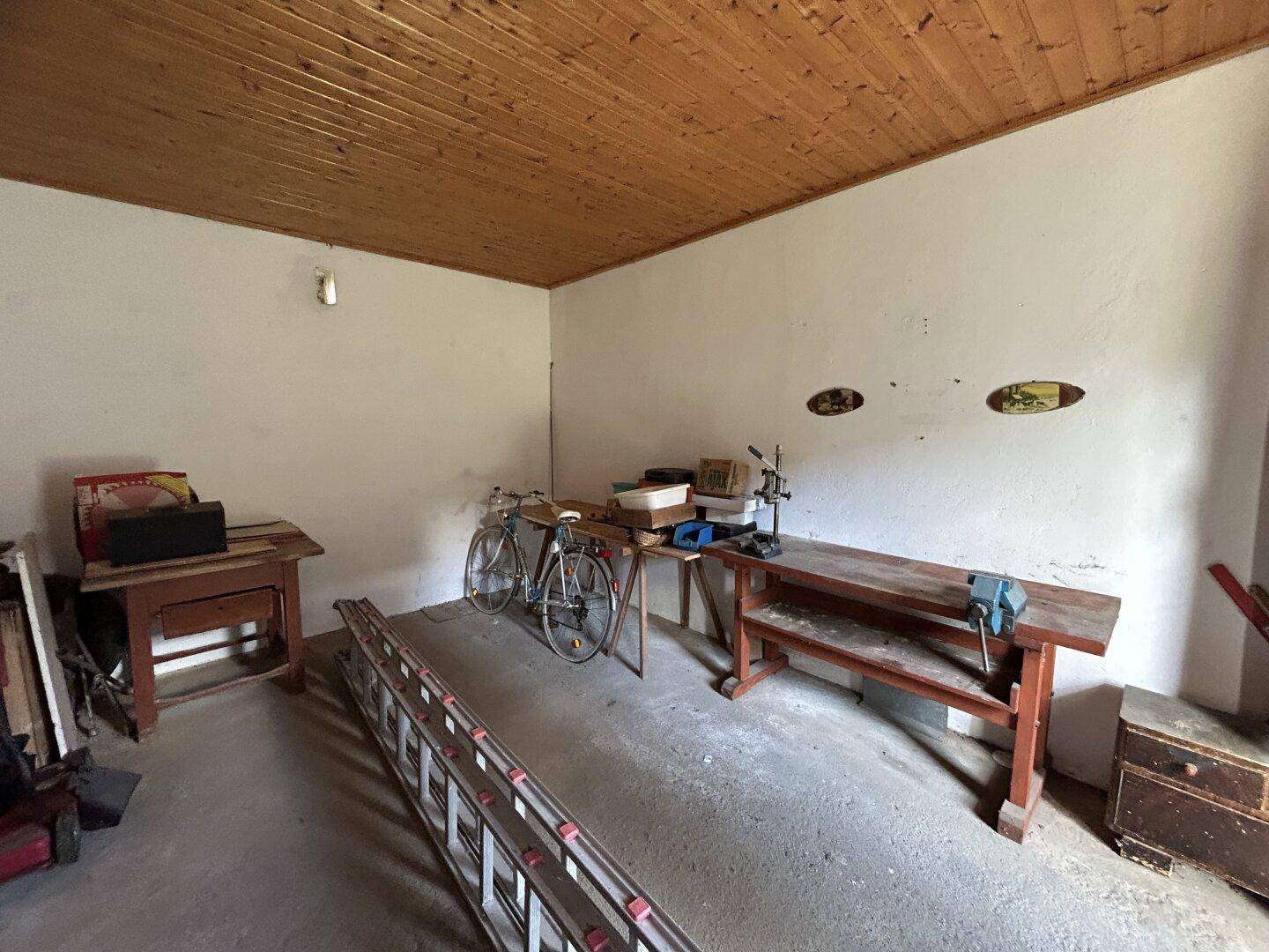 Werkstatt mit Holzbalkendecke, Betonboden und verschiedenen Werkbänken und Geräten.