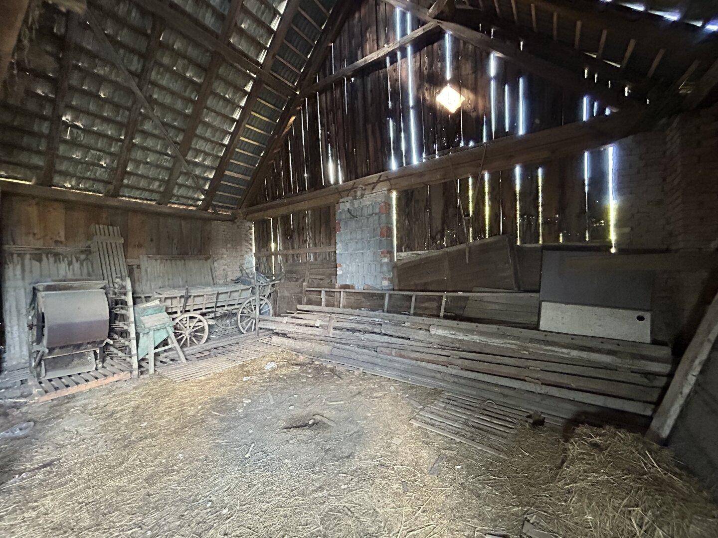 Innenansicht einer Scheune mit Holzbalken, Heu auf dem Boden und alten landwirtschaftlichen Geräten.