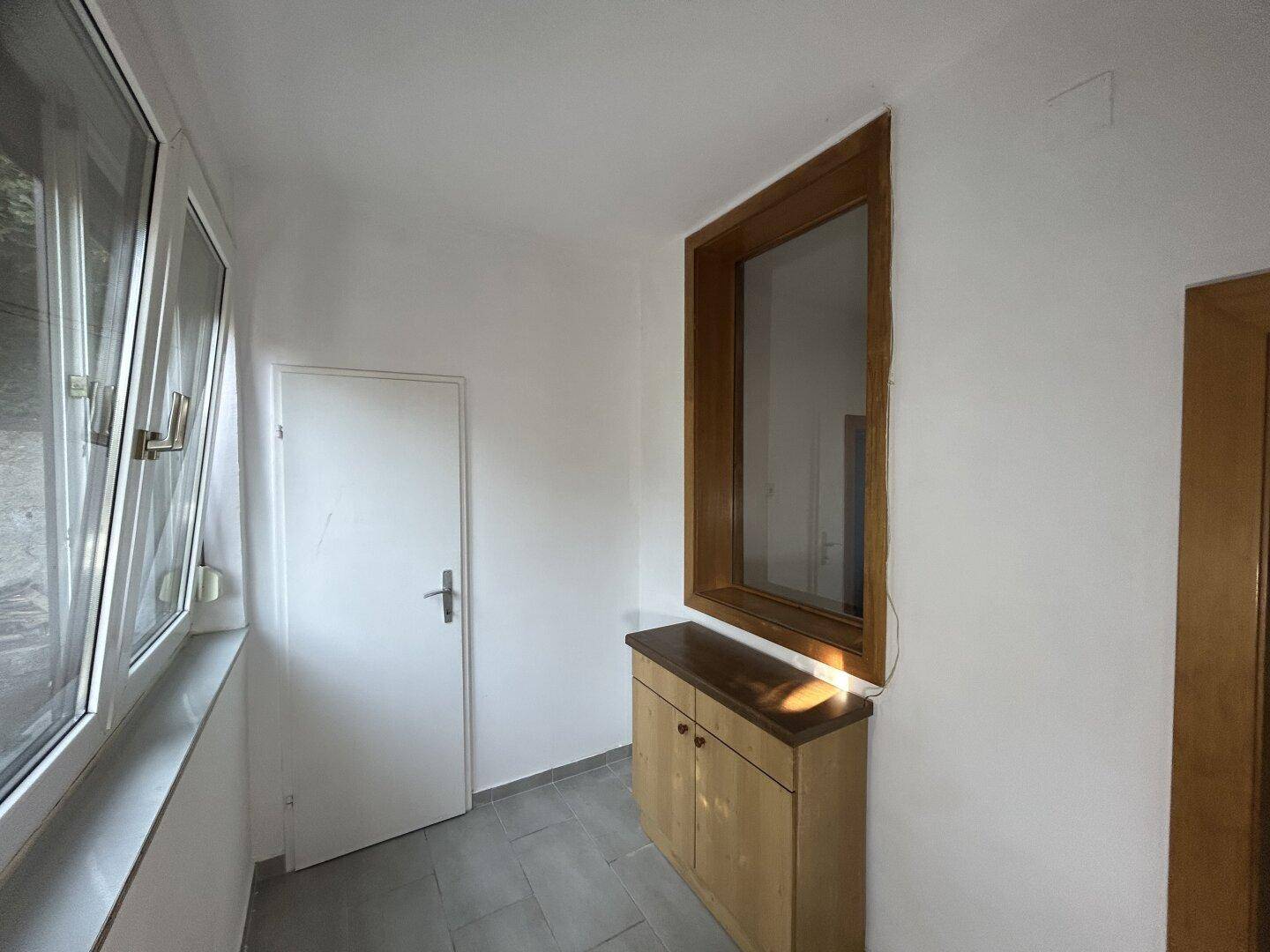 Heller Eingangsbereich mit Fliesenboden, Holzschrank und großen Fenstern für viel Tageslicht.