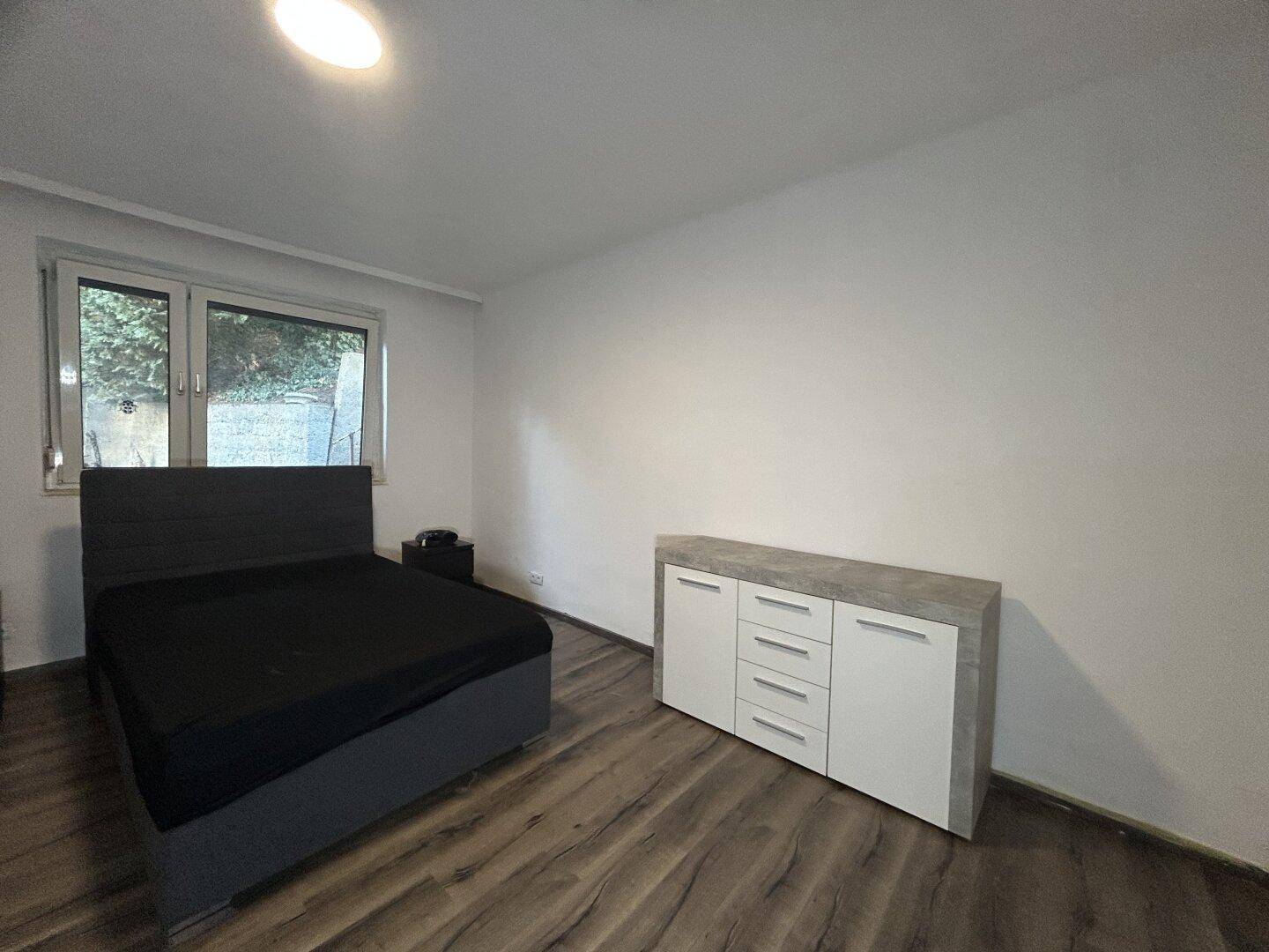 Helles Schlafzimmer mit Bett, Kommode, Laminatboden und Fenster mit Blick ins Grüne.