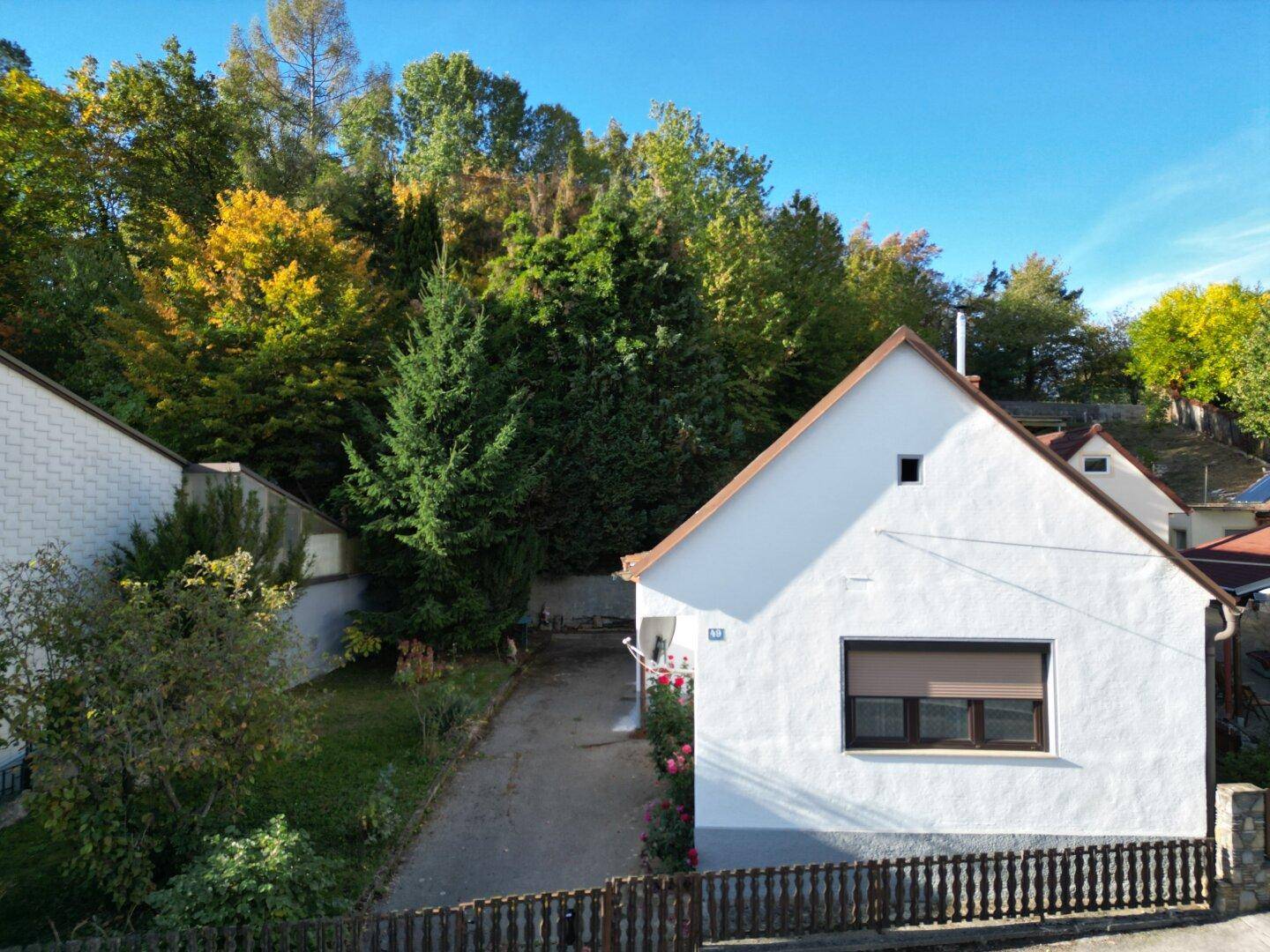Vorderansicht des Hauses mit weißer Fassade, Satteldach und einem kleinen Rasenbereich im Sonnenschein.