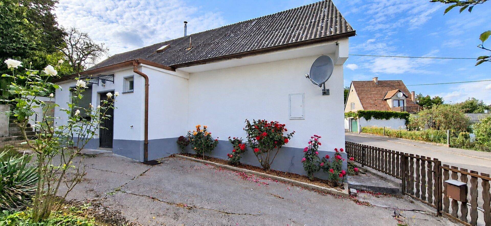 Seitenansicht des Hauses mit blühenden Rosenbüschen und einem gepflasterten Zugang unter blauem Himmel.