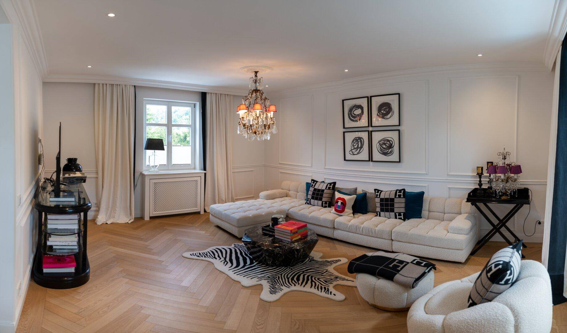 Geräumiges Wohnzimmer mit weißem Sofa, Fischgrätparkett, opulentem Kronleuchter und Stuckdecke.