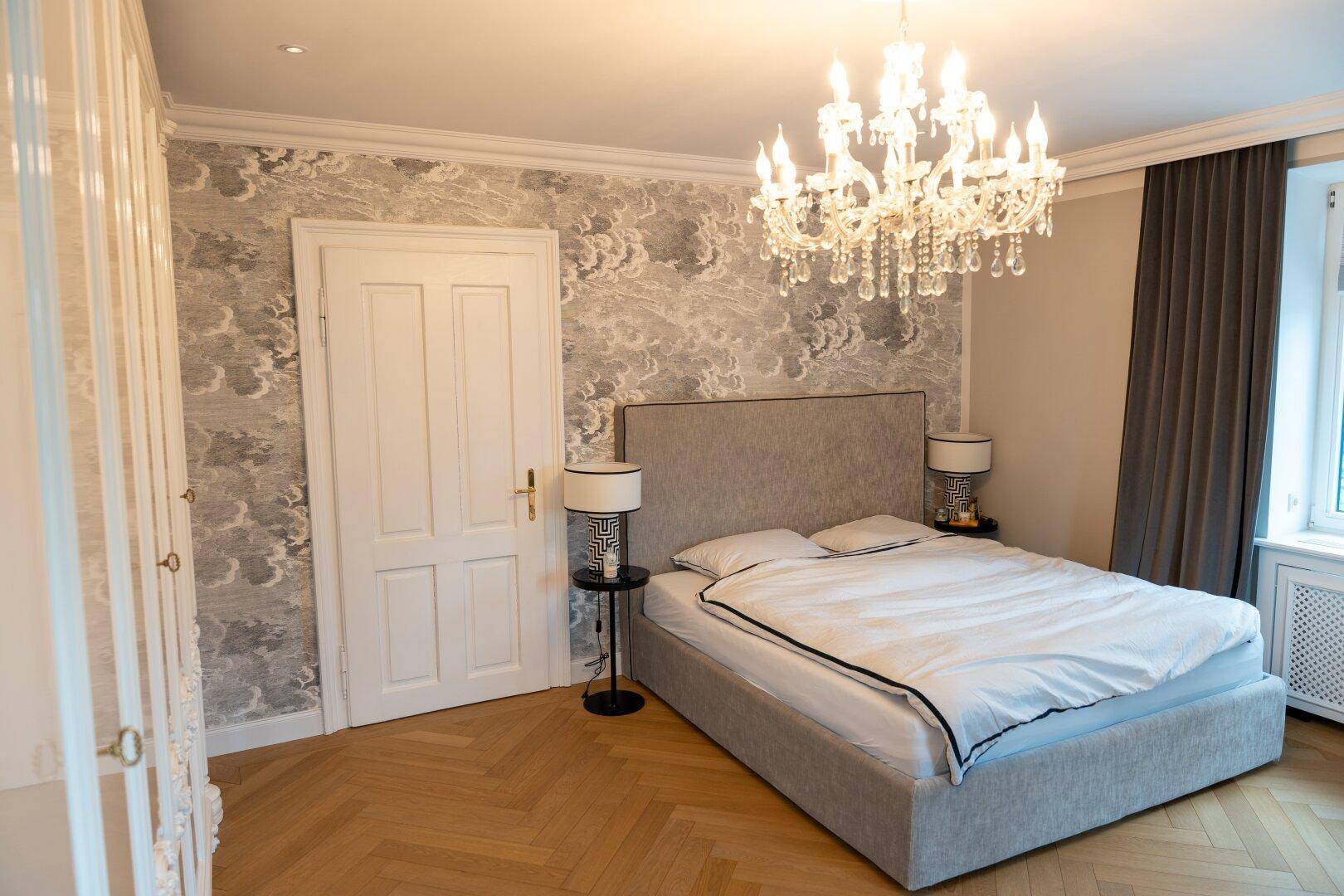 Gemütliches Schlafzimmer mit Doppelbett, gemusterter Tapete und elegantem Kronleuchter, viel Tageslicht.