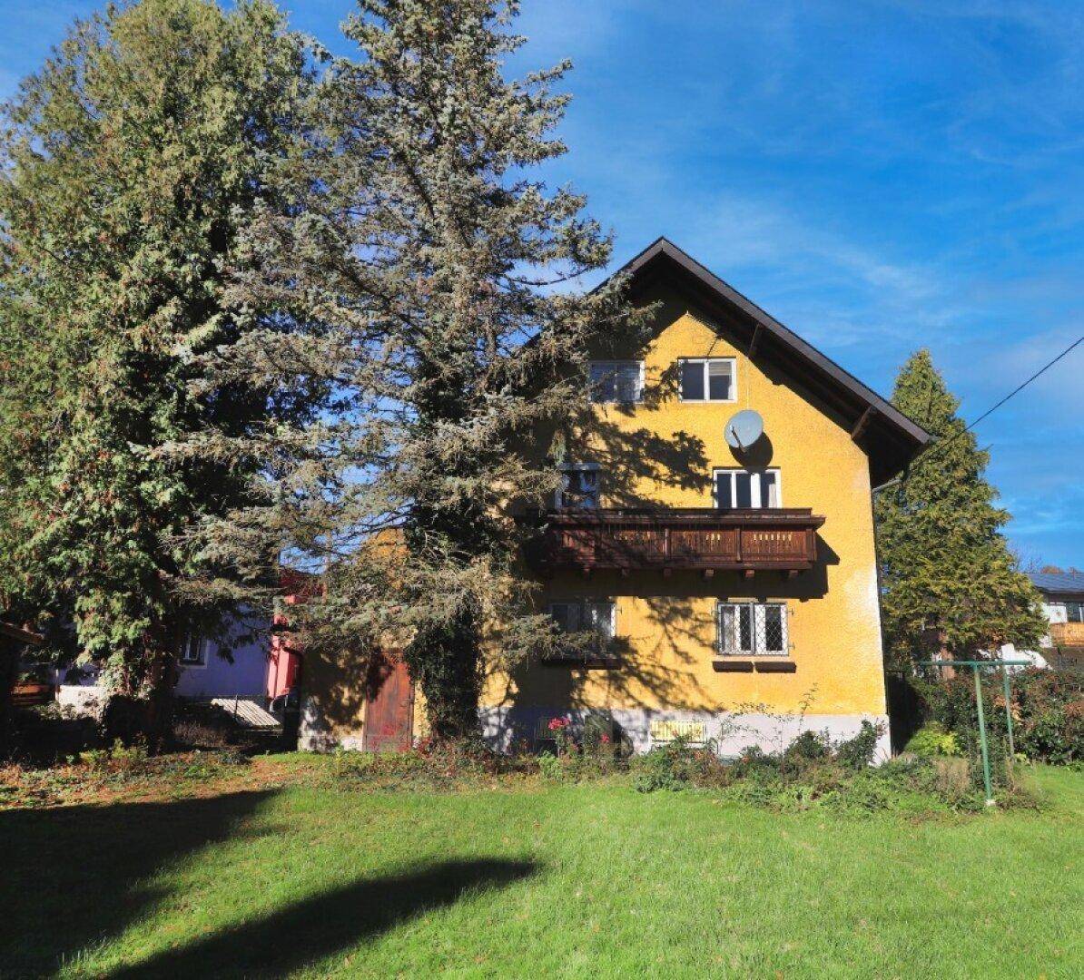 Gelbes Haus mit Holzbalkon und Satteldach, umgeben von hohen Bäumen unter blauem Himmel.