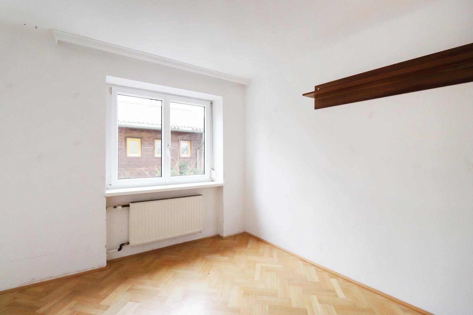 Leeres Zimmer mit hellem Fischgrätparkett, einem Fenster und einfacher Wandgestaltung.
