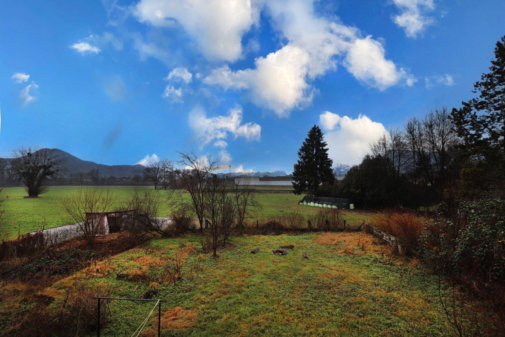 Weitläufige grüne Landschaft mit Feldern, Bäumen und Bergen unter einem blauen Himmel.