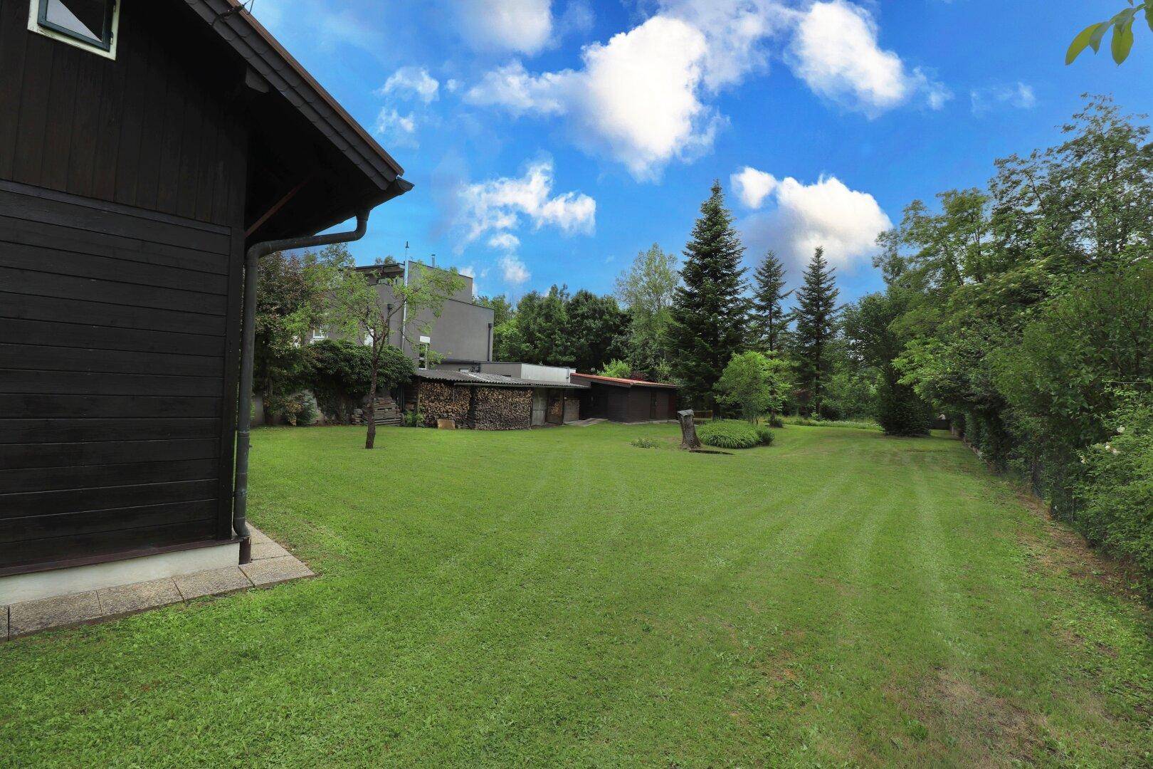 Weitläufiger Garten mit Rasenfläche und dunklem Holzhaus, umgeben von Bäumen unter blauem Himmel.