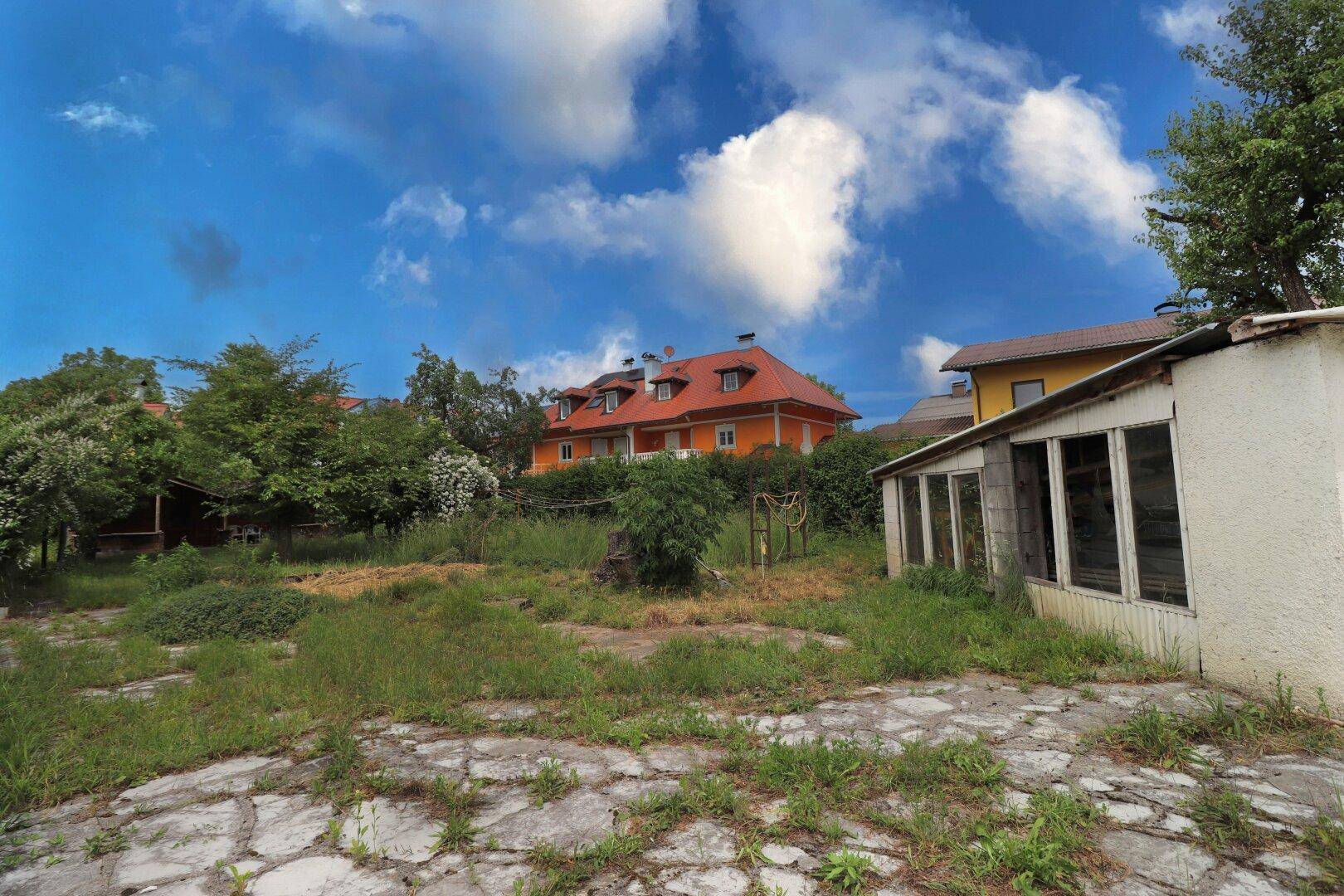 Weitläufiger, ungepflegter Gartenbereich mit einem orangefarbenen Wohnhaus im Hintergrund unter blauem Himmel.