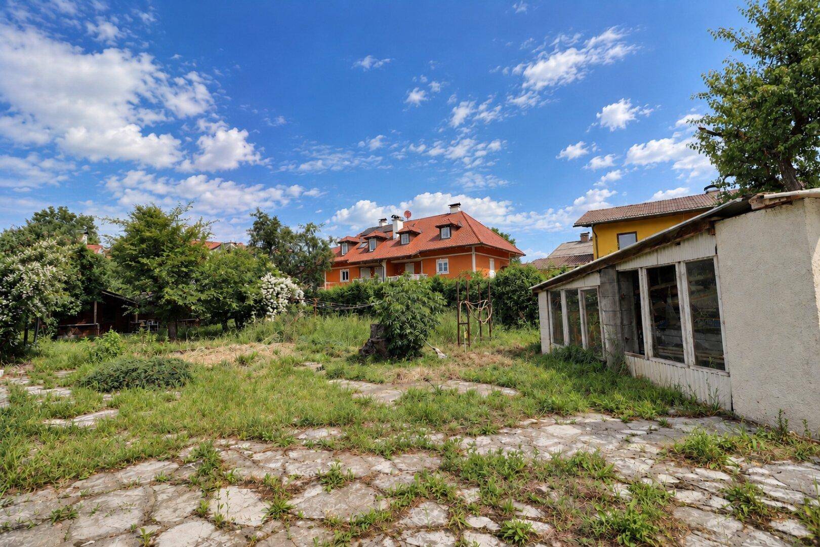 Weitläufiger, verwilderter Gartenbereich mit einer Betonfläche und weiteren Häusern im Hintergrund.