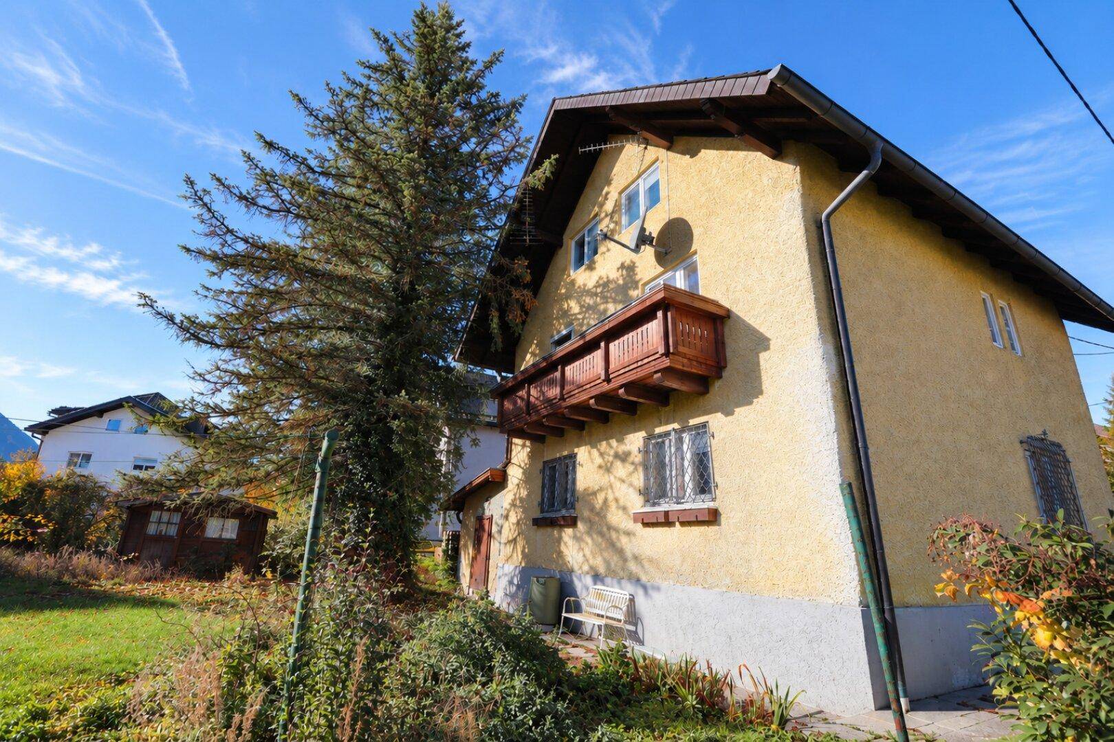Seitenansicht des gelben Wohnhauses mit Holzbalkon und großem Baum im Vordergrund, unter klarem Himmel.