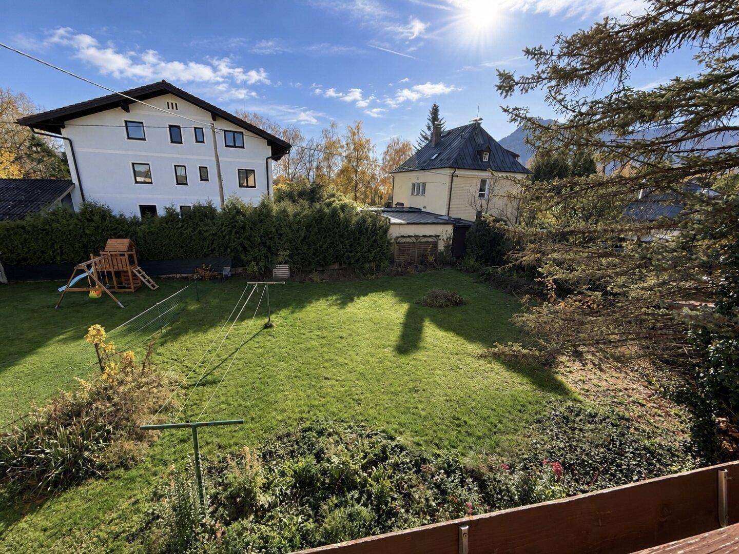 Weitläufiger grüner Garten mit Kinderspielplatz und Hecke, umgeben von weiteren Wohnhäusern bei Sonnenschein.